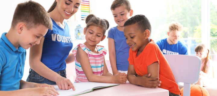 Young volunteer reading book with children at table indoors.