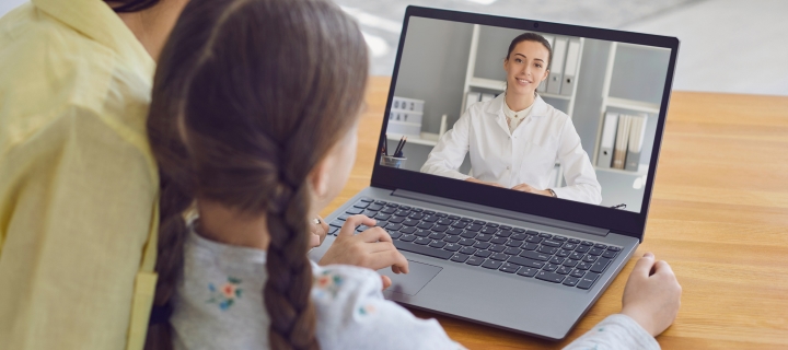 Mother and young daughter consults a doctor with telemedicine using a laptop while sitting at home on the couch.