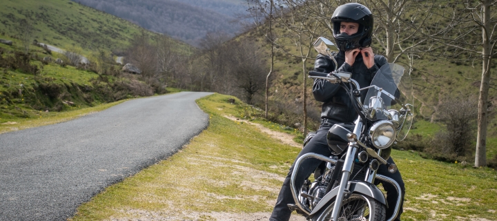 Young biker putting his black helmet on next to a mountain road, sitting on a black custom motorcycle.