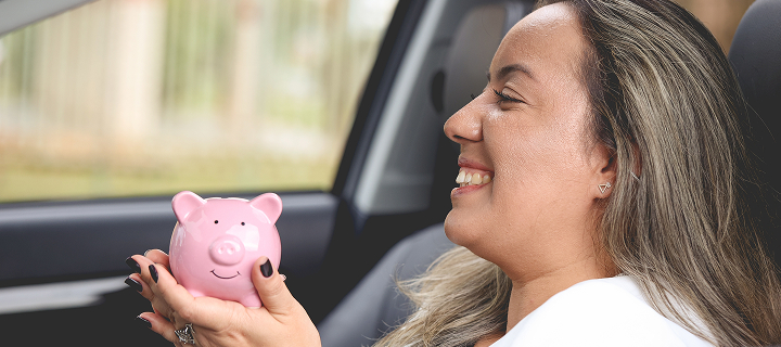A woman sitting in her car, smiling while holding a piggy bank in her hands, saving with car insurance discounts in California through Cost-U-Less. Cheap car insurance in California.