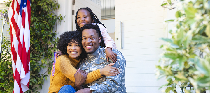 Portrait of a happy African American family hugging outside their home with the American flag; they received a military auto insurance discount with Cost-U-Less. Cheap car insurance in California.