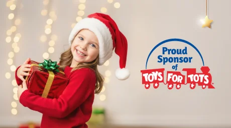 A smiling girl wearing a Santa hat and red sweater holds a wrapped gift in front of holiday lights next to text that reads “Proud Sponsor of Toys for Tots